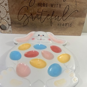 May include: A white ceramic Easter bunny-shaped egg holder with colorful egg-shaped indentations. The bunny has pink ears and a smiling face. A wooden cutting board with the words "Gather Here With Grateful Hearts" is in the background.