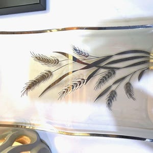 Könnte beinhalten: Rechteckige Servierplatte aus Glas mit weißem Innenraum und silber-goldener Umrandung. Die Platte zeigt ein Weizenähren-Design in Gold und Schwarz. Die Platte wird wahrscheinlich zum Servieren von Speisen oder zum Präsentieren von Dekorationsartikeln verwendet.