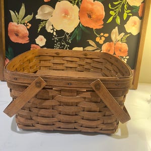 May include: A rectangular woven wooden basket with a medium brown finish, featuring a flat base, vertical sides, and two hinged wooden handles attached to the rim with small metal rivets, showcasing a traditional splint-style construction.