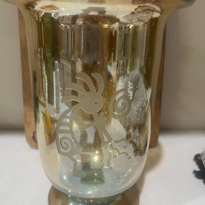 May include: A clear glass candle holder with a flared top and a base. The candle holder has a gold and iridescent finish. A Kokopelli figure is etched on the front. The candle holder is approximately 6 inches tall.