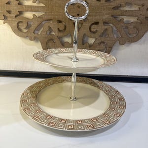 May include: A two-tiered serving stand featuring two round white ceramic plates with a decorative brown and grey geometric Greek key pattern along the rims. The plates are connected by a central polished silver-tone metal rod with a ring handle.