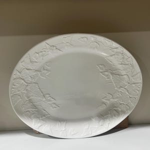 May include: A white, oval-shaped serving platter with a raised leaf design around the rim. The platter is made of ceramic and has a smooth, glossy finish. The leaf pattern adds a touch of elegance to the simple design.