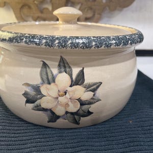 Może przedstawiać: Okrągła, kremowa miska ceramiczna z pokrywką, ozdobiona centralną ilustracją białego kwiatu magnolii z ciemnozielonymi liśćmi. Krawędź miski oraz brzeg pokrywki wykończone są wzorem w ciemnoszare i białe drobne plamki.