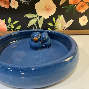 Puede incluir: Un cuenco redondo de cerámica azul intenso que presenta dos pequeños pájaros azules esculpidos con picos marrones unidos al centro de la base interior. El cuenco tiene un acabado liso y brillante y un borde grueso y redondeado.