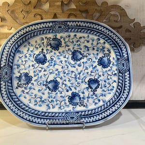Puede incluir: Una bandeja de servicio ovalada de cerámica con base blanca y un patrón floral azul. El diseño incluye hojas azules estilizadas y motivos en forma de corazón, rodeados por un borde geométrico azul y blanco en el borde.