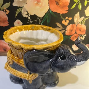 May include: A ceramic planter shaped like a gray elephant with a glossy finish. The elephant features a raised trunk and carries a yellow, basket-weave textured container on its back with a white interior. The design includes small white tusks.