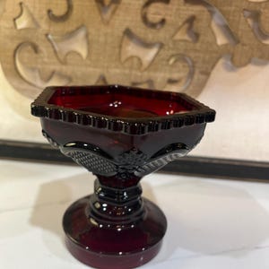 May include: A deep red glass compote dish featuring a hexagonal rim with a scalloped edge. The bowl displays a pressed diamond and fan pattern on its exterior, supported by a thick, multi-tiered pedestal base that rests on a circular foot.