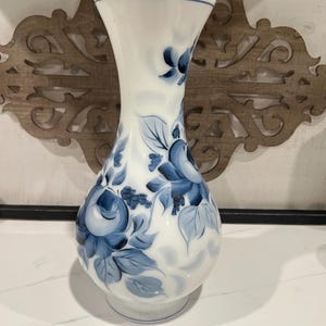 Puede incluir: Un jarrón de vidrio blanco con borde acampanado y cuerpo redondeado, que presenta motivos florales azules pintados a mano con hojas y flores. El diseño incluye varios tonos de azul aplicados con pinceladas sobre la superficie lisa.