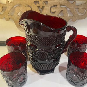Puede incluir: Una jarra de cristal rojo intenso con un patrón de diamantes texturizado y una banda central de motivos circulares, acompañada de cuatro vasos cortos a juego que presentan un patrón similar de diamantes y estrellas.