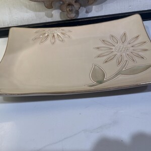 May include: A rectangular beige ceramic tray with slightly curved edges, featuring a large embossed sunflower design on the right side and a smaller partial sunflower motif in the upper left corner, finished with a subtle brown rim.