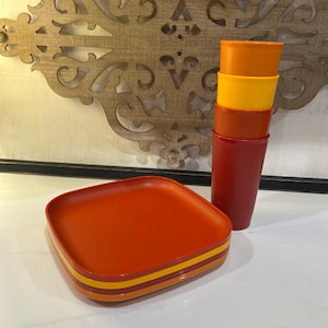 Può includere: Un set di tre piatti quadrati nelle tonalità arancione, giallo e rosso, impilati insieme. Accanto ai piatti c'è una pila di quattro bicchieri nei colori arancione, giallo e rosso coordinati. Gli articoli hanno una finitura opaca e liscia.