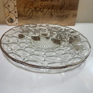 May include: A clear glass plate with a silver rim and floral design. The plate has a diamond pattern and is set against a wooden cutting board with the text "Gather Here With Grateful Hearts."