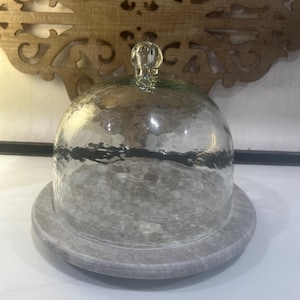 Op de afbeelding: Een heldere glazen stolp met een gehamerde textuur en een kleine bolvormige handgreep, rustend op een ronde grijze marmeren voet. De koepelvormige deksel is transparant, waardoor het gladde stenen oppervlak onder het glas zichtbaar is.