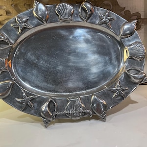 Può includere: Un vassoio da portata ovale in metallo color argento con un bordo rialzato decorato con vari elementi marini in rilievo, tra cui diversi tipi di conchiglie e stelle marine disposte attorno alla superficie centrale liscia e riflettente.