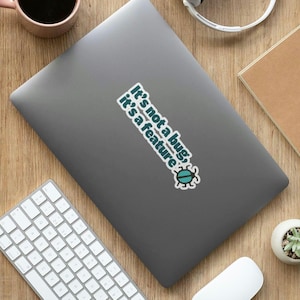 以下が含まれることがあります： 「It's not a bug, it's a feature」と書かれたステッカーが付いたグレーのノートパソコン。漫画の虫のグラフィックも描かれています。白いキーボード、マウス、ヘッドフォン、コーヒーカップも写っています。