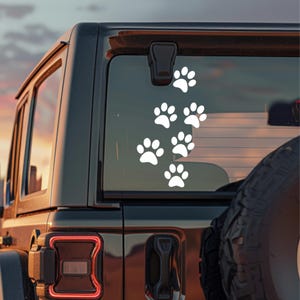 Op de afbeelding: Een witte vinylsticker met een spoor van zes pootafdrukken in een verticaal patroon, aangebracht op de achterruit van een donker voertuig. Het ontwerp bestaat uit effen witte vormen die dierensporen op het glas weergeven.