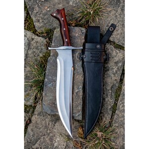May include: A large, silver blade knife with a dark wooden handle and a black leather sheath. The knife has a wide blade and a curved edge. The handle is dark brown with silver rivets.
