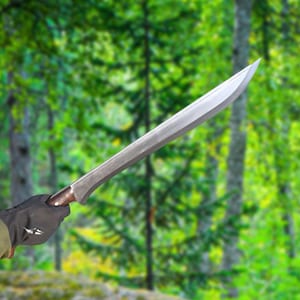 May include: A large, silver machete with a curved blade and a wooden handle. The machete is held by a gloved hand against a backdrop of a blurred green forest. The blade appears sharp and ready for use.
