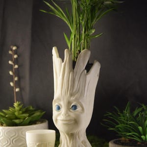 Puede incluir: Una maceta blanca con forma de Groot con ojos azules, que contiene una planta verde. La maceta tiene una cara sonriente y una superficie texturizada. Una pequeña taza blanca con la palabra "NEXTIS" está frente a la maceta. Otras plantas están en el fondo.