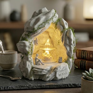 May include: A gray stone-like sculpture of a cave with a glowing candle inside. The sculpture has a small figure of a person sitting on a ledge. Green vines and the word "VERTIS" are visible.