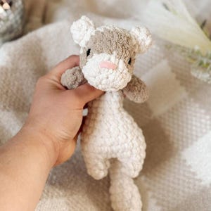 May include: A handmade crochet cat plushie in white, gray, and pink. The cat has a textured, soft appearance with a small pink nose and gray patches on its head. The plush toy is held against a soft, textured background.