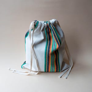 Peut inclure: Un sac à cordon blanc avec un motif à rayures colorées vertes, oranges, jaunes et bleues. Le sac a un design simple avec une fermeture à cordon et est fait d'un tissu robuste. Le sac convient au transport de divers articles.