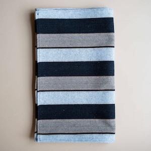 May include: A folded rectangular cloth with horizontal stripes in black, grey, and light blue. The cloth appears to be made of woven fabric, with visible texture. The stripes are evenly spaced across the surface. The edges are neatly finished.