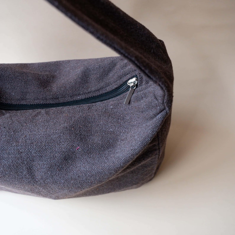 May include: A dark gray crossbody bag with a zipper closure and a matching strap. The bag is made of a textured fabric and has a silver zipper pull. The strap is wide and appears to be made of the same material as the bag.