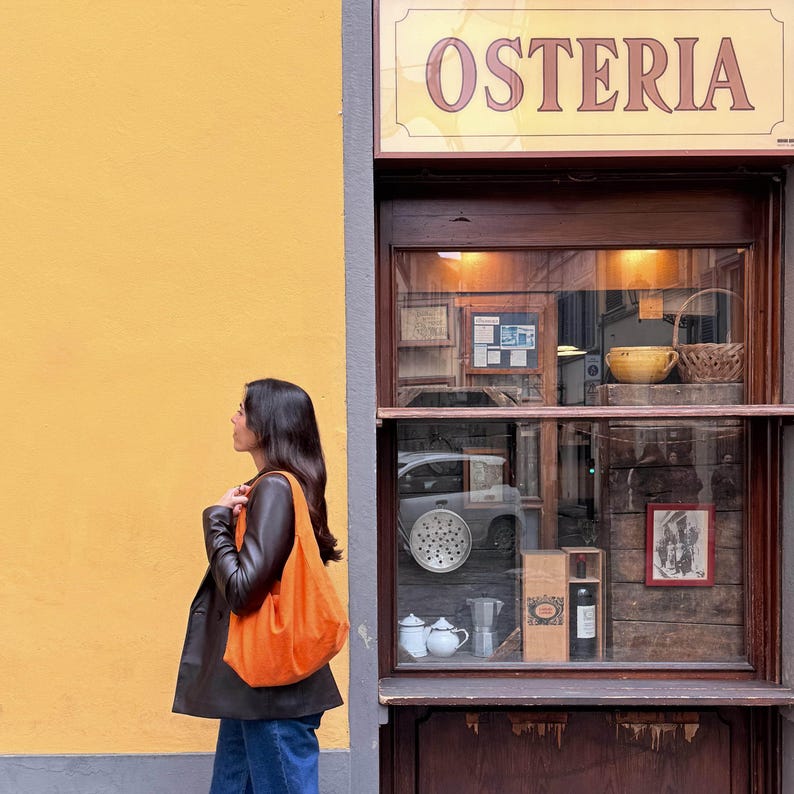 May include: An orange tote bag is carried by a person walking past a yellow wall and a storefront with the word "OSTERIA" above the window. The storefront has a wooden frame and displays various items inside.