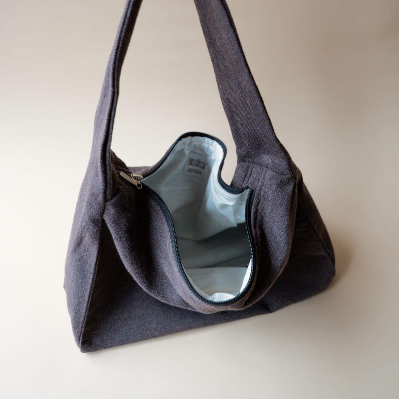 May include: A dark gray shoulder bag with a zipper closure. The bag has a wide strap and a light blue interior. The bag is made of a textured fabric and is open, revealing the interior.