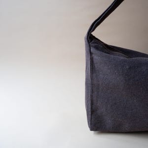 May include: A dark gray shoulder bag made of a textured fabric. The bag has a single strap and a rectangular shape. The bag is shown against a light gray background. The bag is a simple, minimalist design.