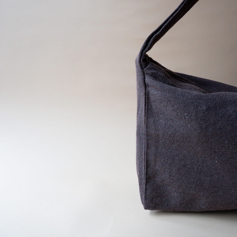 May include: A dark gray shoulder bag made of a textured fabric. The bag has a single strap and a rectangular shape. The bag is shown against a light gray background. The bag is a simple, minimalist design.