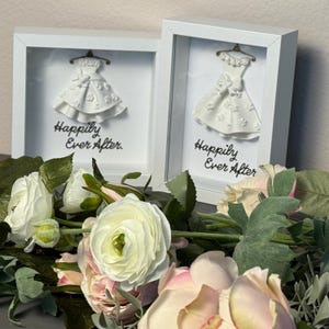 Puede incluir: Dos marcos tipo caja de sombra blancos que contienen vestidos blancos en miniatura en perchas con detalles florales y el texto Happily Ever After impreso en negro. Los marcos están colocados detrás de flores artificiales blancas y rosa pálido.