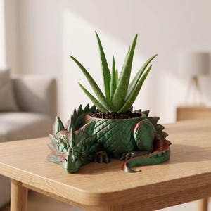 May include: A green dragon-shaped planter with a metallic finish, featuring detailed scales and a curled tail. The dragon's back forms a circular container holding a green aloe vera plant with long, pointed leaves planted in dark soil.
