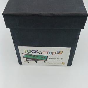 May include: A black square box with a lid. The top of the lid features the gold text "Objet d'art" and a small logo. A white rectangular label on the front displays the text "rackem'up" with a graphic of a pool table, "Release No. 181", and "Objet d'art".