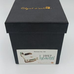 May include: A square black box with a lid featuring the gold text Objet d'art. A white rectangular label on the front displays the text Release No. 158, 1957 heaven!, and a small illustration of a vintage car with teal and white paint.