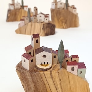 Può includere: Tre sculture di villaggio in miniatura realizzate con blocchi di legno naturale. Ogni pezzo presenta piccole case con tetti rossi, una chiesa centrale con una croce e un singolo albero verde, montati su una base in legno chiaro e strutturato.