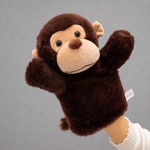May include: A brown plush monkey hand puppet with a tan face, ears, and muzzle. The puppet has large black eyes and is being worn on a hand. The puppet's arms are raised, and a small tag is visible.