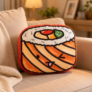 Puede incluir: Un cojín de peluche con forma de rollo de sushi con un exterior color salmón que presenta una cara de dibujos animados con un ojo guiñando y una pequeña lengua. La superficie superior muestra arroz blanco, un centro rojo y una sección verde.