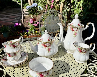 Könnte beinhalten: Ein Teeservice aus weißem Porzellan mit einem zarten Blumenmuster aus roten und rosafarbenen Rosen, grünen Blättern und Goldrand. Das Set besteht aus einer hohen Teekanne, einer kleineren Teekanne, einem Milchkännchen, einer Zuckerdose und zwei Tassen.