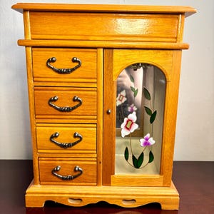 May include: A wooden jewellery box with four drawers on the left and a glass-door cabinet on the right. The cabinet door features a floral design with white, pink, and purple flowers. The box has a light brown finish and ornate drawer pulls.
