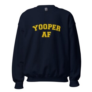 Può includere: Una felpa blu navy con la scritta "YOOPER AF" in lettere gialle. La felpa ha un girocollo e maniche lunghe. Il testo è in un font in stile collegiale.