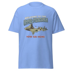 Può includere: T-shirt azzurra con una grafica della Penisola Superiore del Michigan. La grafica include il testo "WELCOME TO THE UPPER PENINSULA" in verde e "NOW GO HOME" in rosso, insieme a illustrazioni di punti di riferimento.
