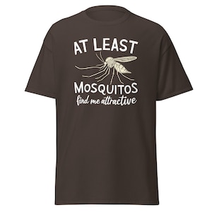 May include: A brown t-shirt with the text "AT LEAST MOSQUITOS find me attractive" in white. A detailed illustration of a mosquito is above the text. The shirt is a short-sleeved, casual style.