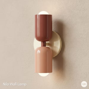 May include: A contemporary wall lamp featuring a two-tone colour scheme. The top section is a deep reddish-brown, and the bottom section is a soft peach. It has a cream-coloured circular base and emits a warm light from two spherical bulbs. The text "Nilo Wall Lamp" is at the bottom.