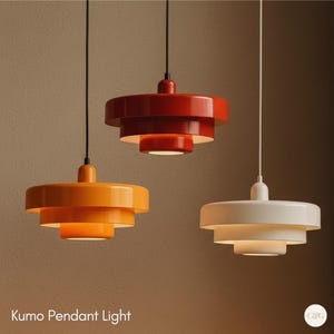 May include: Three modern pendant lights in the image. One is orange, one is red, and one is white. Each light has a tiered, disc-shaped design. The lights hang from black or white cords. The text "Kumo Pendant Light" is at the bottom.