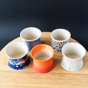 Op de afbeelding: Vijf handgemaakte keramische bekers met een uitlopend ontwerp. De bekers hebben een verscheidenheid aan kleuren en patronen, waaronder blauw-witte wervelingen, een gespikkeld patroon en een oranje verloop. De binnenkanten zijn wit en de bekers worden op een houten oppervlak gepresenteerd.
