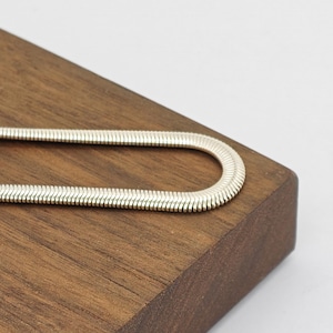 May include: A silver-toned snake chain necklace with a flat, flexible, and tightly woven link design. The chain is bent into a U-shape, showcasing its smooth, metallic surface and uniform, interlocking pattern that creates a continuous, sleek appearance.