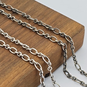Puede incluir: Dos cadenas de metal plateado con eslabones ovalados. Las cadenas están dispuestas sobre un bloque de madera, mostrando un acabado pulido en la cadena frontal y un acabado oxidado más oscuro en la cadena situada detrás.