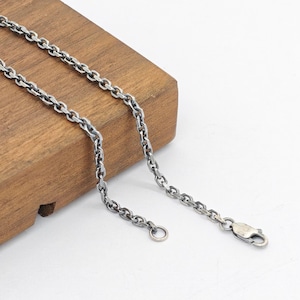 Puede incluir: Un collar de cadena de metal plateado con eslabones ovalados y un acabado oscuro y oxidado. La cadena incluye un cierre de mosquetón en un extremo y una pequeña anilla circular en el otro, mostrando una apariencia texturizada e industrial.